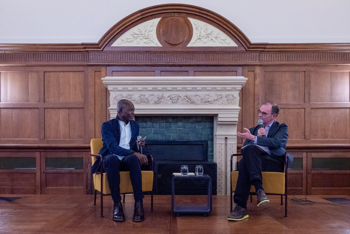Two men seated in armchairs arranged as for an interview in a grand hall converse.